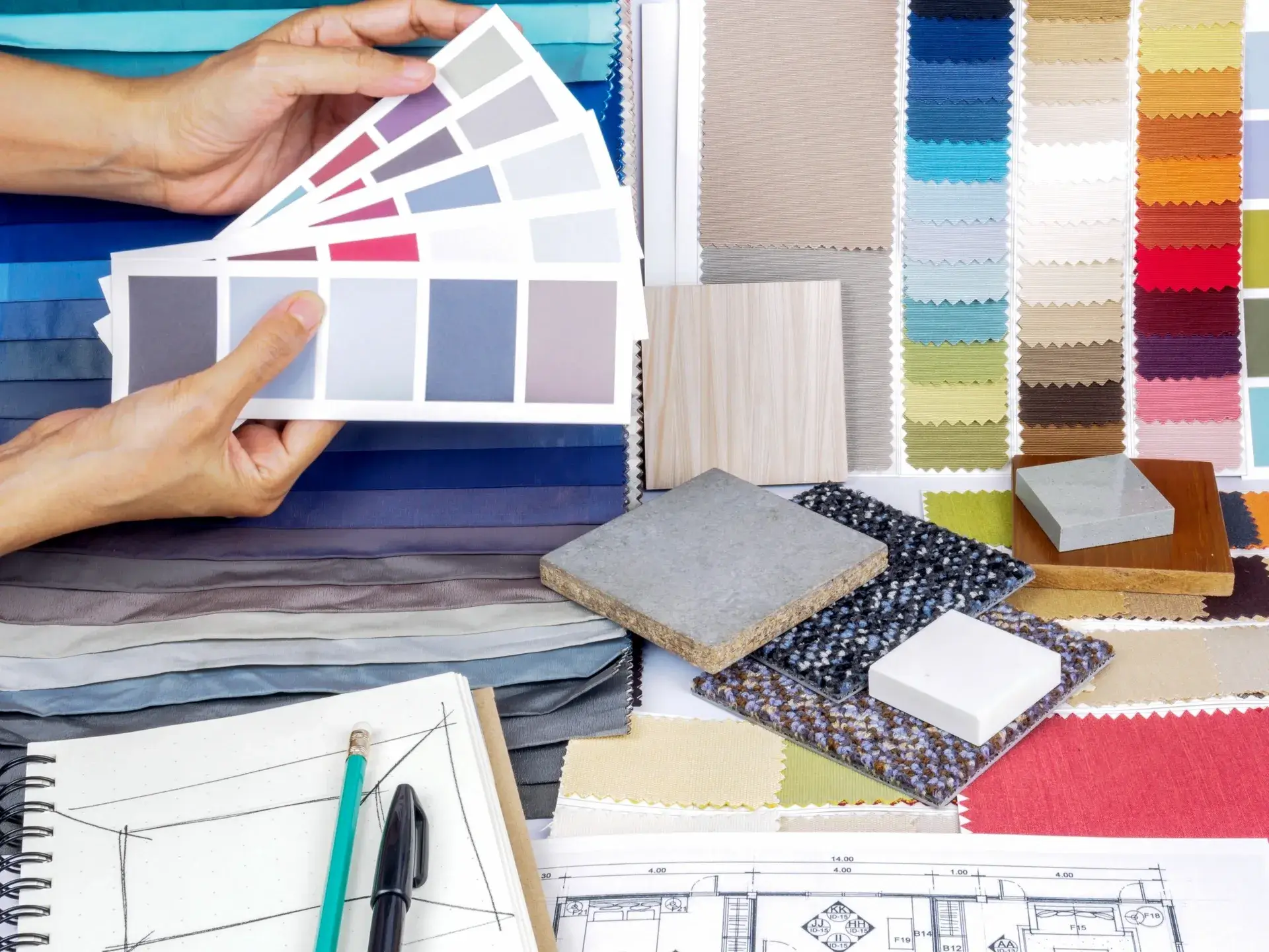 Hands holding a color swatch book over fabric samples and design plans.